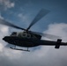 A military helicopter is captured in flight against a partly cloudy sky, photographed from a low angle looking upward. The aircraft appears to be a dark olive/green colored utility helicopter, likely a Bell 412 or similar variant, with rotor blades in motion creating visible motion blur. The image has a dramatic, low-angle perspective emphasizing the helicopter's silhouette against the overcast sky.