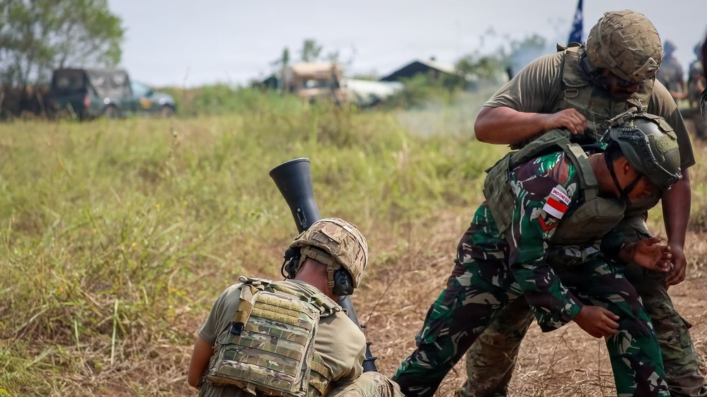 The image depicts what appears to be a military training exercise involving soldiers from at least two different nations. One soldier wearing Indonesian military camouflage (identifiable by the red-and-white flag patch) is being assisted or restrained by another soldier in olive drab uniform. A third soldier in multicam pattern gear is positioned in the foreground with what appears to be a grenade launcher attached to a rifle.