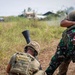 The image depicts what appears to be a military training exercise involving soldiers from at least two different nations. One soldier wearing Indonesian military camouflage (identifiable by the red-and-white flag patch) is being assisted or restrained by another soldier in olive drab uniform. A third soldier in multicam pattern gear is positioned in the foreground with what appears to be a grenade launcher attached to a rifle.