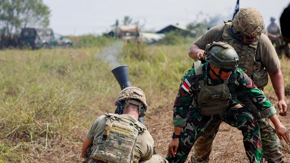 The image shows a joint military training exercise involving soldiers in two distinct uniforms — multicam/OCP pattern and Indonesian camouflage pattern. A soldier in the foreground is firing or handling a mortar tube, with smoke visible at the muzzle. Other personnel are observing and assisting in close proximity on an open grassy field.