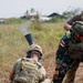 The image shows a joint military training exercise involving soldiers in two distinct uniforms — multicam/OCP pattern and Indonesian camouflage pattern. A soldier in the foreground is firing or handling a mortar tube, with smoke visible at the muzzle. Other personnel are observing and assisting in close proximity on an open grassy field.