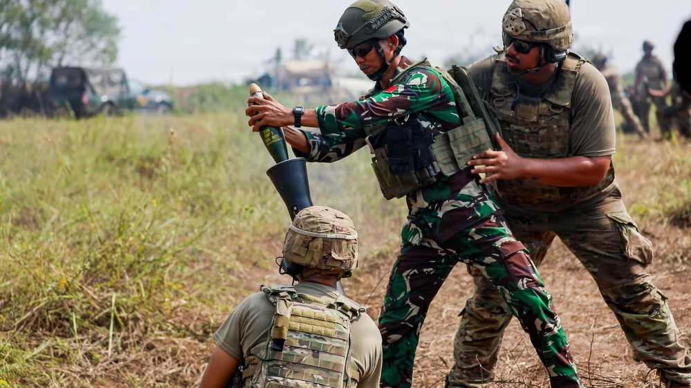 Military personnel in mixed uniforms are conducting a mortar training exercise in an open field. One soldier in Indonesian camouflage pattern (TNI) is loading or demonstrating a mortar round into a mortar tube, assisted by soldiers in multicam/OCP uniforms. The scene appears to be a joint military training exercise based on the mixed uniform types present.