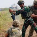 Military personnel in mixed uniforms are conducting a mortar training exercise in an open field. One soldier in Indonesian camouflage pattern (TNI) is loading or demonstrating a mortar round into a mortar tube, assisted by soldiers in multicam/OCP uniforms. The scene appears to be a joint military training exercise based on the mixed uniform types present.