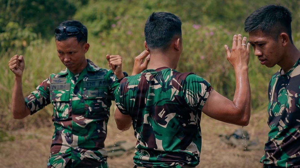 Three military personnel wearing matching green camouflage uniforms are engaged in what appears to be a hand-to-hand combat or martial arts training exercise outdoors. The central figure, whose back is to the camera, appears wet and is in a defensive or striking stance facing a third soldier on the right. A fourth soldier on the left is also in a fighting stance, observing or participating in the drill.