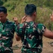 Three military personnel wearing matching green camouflage uniforms are engaged in what appears to be a hand-to-hand combat or martial arts training exercise outdoors. The central figure, whose back is to the camera, appears wet and is in a defensive or striking stance facing a third soldier on the right. A fourth soldier on the left is also in a fighting stance, observing or participating in the drill.