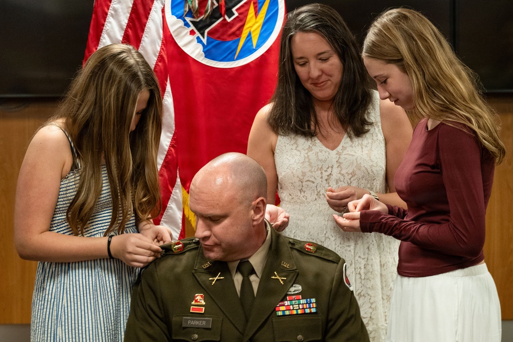 A U.S. Army officer named Parker is seated during what appears to be a military promotion ceremony. Three women, likely family members, are pinning new rank insignia onto his uniform collar and shoulders. The ceremony takes place in front of military flags, including what appears to be the Tennessee state flag and a unit colors flag.