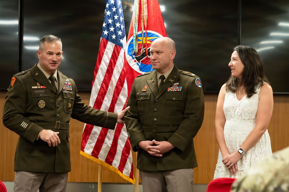 A military ceremony featuring two U.S. Army officers in olive green dress uniforms standing before an American flag and a unit colors flag. A civilian woman in a white lace dress stands to the right, likely a family member participating in the ceremony. The officer on the left, whose name tape reads 'CARPENTER,' has his hand on the shoulder of the center officer in a gesture of recognition or congratulation.