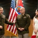A military ceremony featuring two U.S. Army officers in olive green dress uniforms standing before an American flag and a unit colors flag. A civilian woman in a white lace dress stands to the right, likely a family member participating in the ceremony. The officer on the left, whose name tape reads 'CARPENTER,' has his hand on the shoulder of the center officer in a gesture of recognition or congratulation.