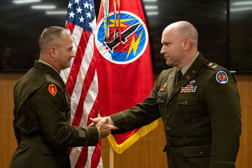 Two U.S. Army officers in dress green uniforms are shaking hands in front of an American flag and a red unit guidon/flag bearing a circular insignia with lightning bolt and missile imagery. The setting appears to be an official military ceremony, likely an award presentation, promotion, or change of command. Both individuals wear unit patches and service ribbons consistent with senior military personnel.