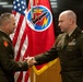 Two U.S. Army officers in dress green uniforms are shaking hands in front of an American flag and a red unit guidon/flag bearing a circular insignia with lightning bolt and missile imagery. The setting appears to be an official military ceremony, likely an award presentation, promotion, or change of command. Both individuals wear unit patches and service ribbons consistent with senior military personnel.