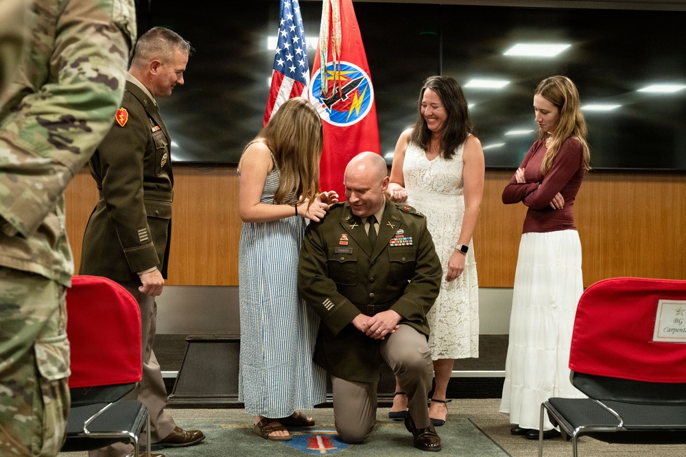 A military promotion or pinning ceremony is taking place indoors, likely at a U.S. Army installation. A uniformed soldier is kneeling while a young woman pins new rank insignia onto his shoulder, surrounded by family members and a senior officer. The setting includes U.S. and unit flags in the background.