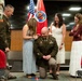 A military promotion or pinning ceremony is taking place indoors, likely at a U.S. Army installation. A uniformed soldier is kneeling while a young woman pins new rank insignia onto his shoulder, surrounded by family members and a senior officer. The setting includes U.S. and unit flags in the background.