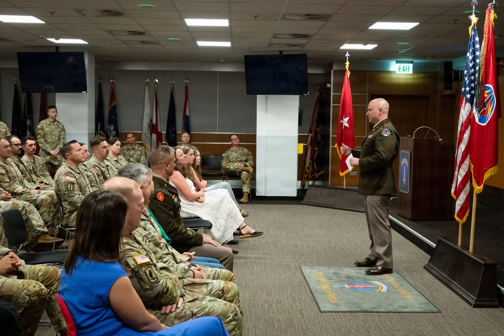 A military ceremony or briefing is taking place in a formal conference or auditorium room. A senior officer in Army Service Uniform (green) is addressing an audience of seated military personnel in camouflage uniforms and a few civilians. The setting includes multiple military flags, a podium, and a unit insignia floor mat.
