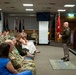 A military ceremony or briefing is taking place in a formal conference or auditorium room. A senior officer in Army Service Uniform (green) is addressing an audience of seated military personnel in camouflage uniforms and a few civilians. The setting includes multiple military flags, a podium, and a unit insignia floor mat.