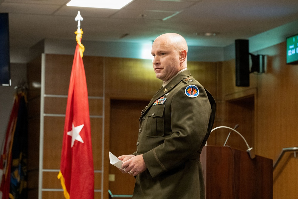 A bald male U.S. Army officer in dress green uniform stands near a podium in what appears to be a formal military briefing or ceremony room. He is holding a small piece of paper and appears to be speaking or preparing to speak. A red general officer flag with a single white star is visible in the background, indicating a one-star (Brigadier General) command presence.