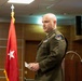 A bald male U.S. Army officer in dress green uniform stands near a podium in what appears to be a formal military briefing or ceremony room. He is holding a small piece of paper and appears to be speaking or preparing to speak. A red general officer flag with a single white star is visible in the background, indicating a one-star (Brigadier General) command presence.