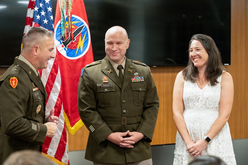 A military ceremony featuring two U.S. Army officers in dress green uniforms and a civilian woman in a white dress. The group appears to be sharing a lighthearted moment, likely during a promotion or award ceremony. An American flag and what appears to be a Tennessee National Guard flag are visible in the background.