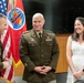 A military ceremony featuring two U.S. Army officers in dress green uniforms and a civilian woman in a white dress. The group appears to be sharing a lighthearted moment, likely during a promotion or award ceremony. An American flag and what appears to be a Tennessee National Guard flag are visible in the background.