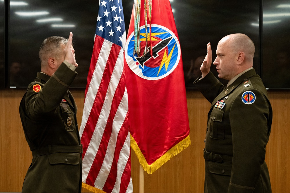 Two U.S. Army officers in dress green uniforms are conducting a formal military oath ceremony, both with right hands raised in salute position. They are standing before an American flag and a red unit guidon/flag bearing a circular insignia with lightning bolt imagery. The setting appears to be an official indoor military facility.