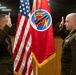 Two U.S. Army officers in dress green uniforms are conducting a formal military oath ceremony, both with right hands raised in salute position. They are standing before an American flag and a red unit guidon/flag bearing a circular insignia with lightning bolt imagery. The setting appears to be an official indoor military facility.
