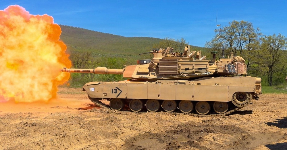 An M1 Abrams main battle tank painted in desert tan is firing its main gun, producing a large orange muzzle blast. The tank is marked with the number '13' and the name 'COOKIE MONSTER' on the gun barrel. The scene takes place on a dirt training range with a forested hillside and blue sky in the background.