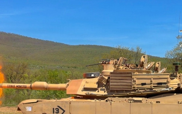 An M1 Abrams main battle tank painted in desert tan is firing its main gun, producing a large orange muzzle blast. The tank is marked with the number '13' and the name 'COOKIE MONSTER' on the gun barrel. The scene takes place on a dirt training range with a forested hillside and blue sky in the background.