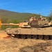 An M1 Abrams main battle tank painted in desert tan is firing its main gun, producing a large orange muzzle blast. The tank is marked with the number '13' and the name 'COOKIE MONSTER' on the gun barrel. The scene takes place on a dirt training range with a forested hillside and blue sky in the background.