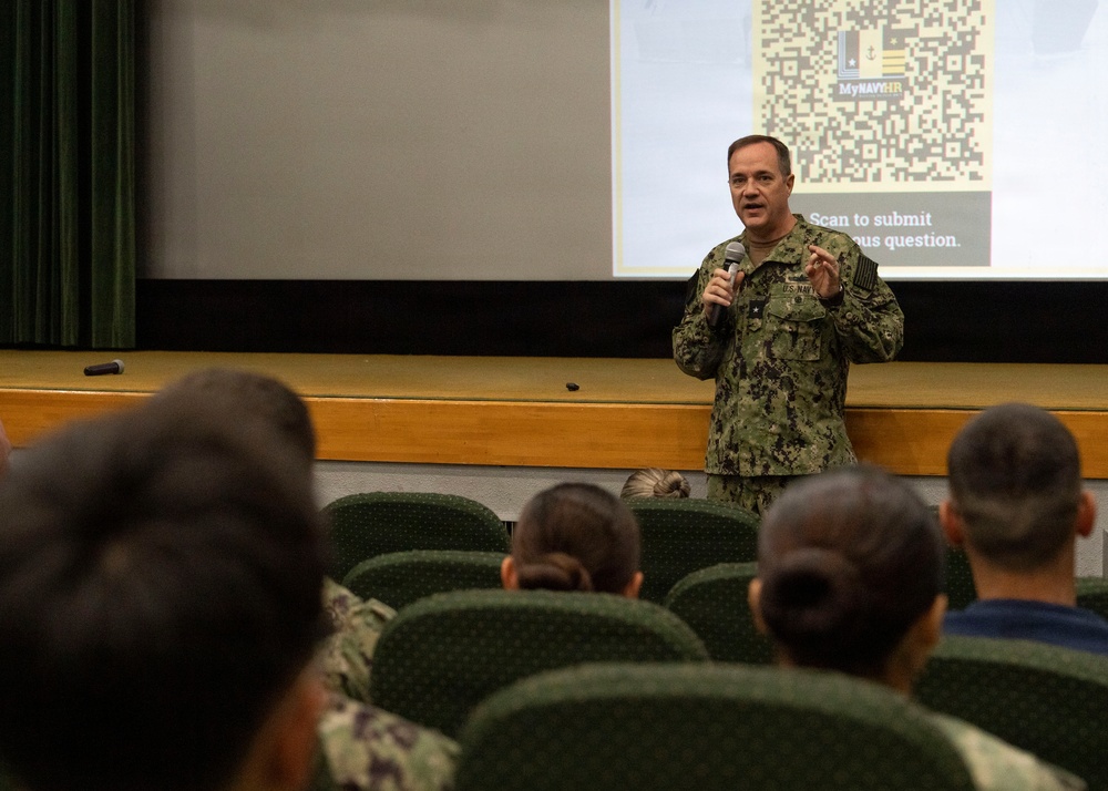 A U.S. Navy officer in NWU Type III (digital camouflage) uniform is addressing an audience in an auditorium or briefing room. He is holding a microphone and speaking in front of a projected presentation slide displaying a QR code and the 'MyNavyHR' branding with text reading 'Scan to submit [anonym]ous question.' The audience, composed of military personnel seated in green chairs, is listening attentively with their backs to the camera.