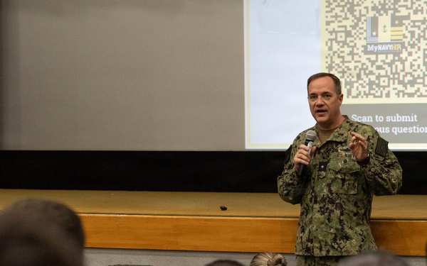 A U.S. Navy officer in NWU Type III (digital camouflage) uniform is addressing an audience in an auditorium or briefing room. He is holding a microphone and speaking in front of a projected presentation slide displaying a QR code and the 'MyNavyHR' branding with text reading 'Scan to submit [anonym]ous question.' The audience, composed of military personnel seated in green chairs, is listening attentively with their backs to the camera.