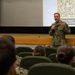 A U.S. Navy officer in NWU Type III (digital camouflage) uniform is addressing an audience in an auditorium or briefing room. He is holding a microphone and speaking in front of a projected presentation slide displaying a QR code and the 'MyNavyHR' branding with text reading 'Scan to submit [anonym]ous question.' The audience, composed of military personnel seated in green chairs, is listening attentively with their backs to the camera.