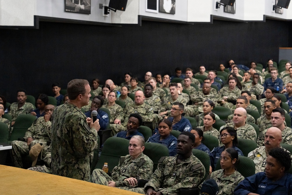 A senior U.S. Navy officer in camouflage NWU (Navy Working Uniform) is addressing a large assembly of military personnel in an auditorium or theater-style venue. The audience consists of dozens of sailors and service members seated in green upholstered chairs, attentively listening to the speaker who is holding a microphone. The setting appears to be an all-hands meeting or command briefing at a military installation.