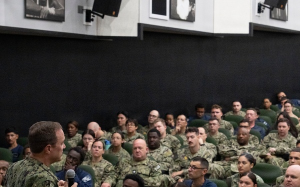 A senior U.S. Navy officer in camouflage NWU (Navy Working Uniform) is addressing a large assembly of military personnel in an auditorium or theater-style venue. The audience consists of dozens of sailors and service members seated in green upholstered chairs, attentively listening to the speaker who is holding a microphone. The setting appears to be an all-hands meeting or command briefing at a military installation.