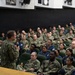 A senior U.S. Navy officer in camouflage NWU (Navy Working Uniform) is addressing a large assembly of military personnel in an auditorium or theater-style venue. The audience consists of dozens of sailors and service members seated in green upholstered chairs, attentively listening to the speaker who is holding a microphone. The setting appears to be an all-hands meeting or command briefing at a military installation.