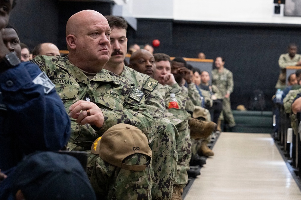 A group of U.S. Navy personnel are seated in rows, appearing to attend a briefing, ceremony, or presentation in an indoor facility. The foreground features a bald male sailor with the name tape 'DEVORAK' and 'U.S. NAVY' tape visible on his uniform, holding a tan cap with 'CO' lettering and gold stars. The assembled personnel are in NWU (Navy Working Uniform) digital camouflage pattern.
