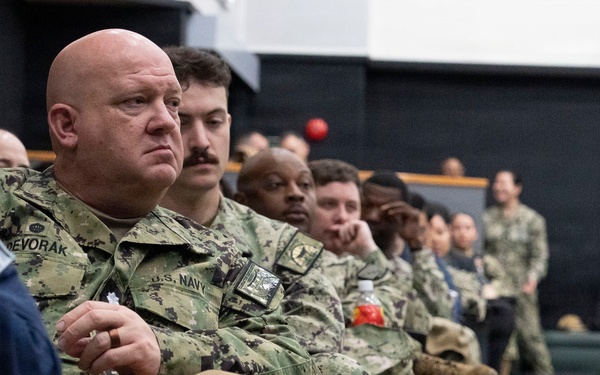 A group of U.S. Navy personnel are seated in rows, appearing to attend a briefing, ceremony, or presentation in an indoor facility. The foreground features a bald male sailor with the name tape 'DEVORAK' and 'U.S. NAVY' tape visible on his uniform, holding a tan cap with 'CO' lettering and gold stars. The assembled personnel are in NWU (Navy Working Uniform) digital camouflage pattern.
