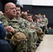 A group of U.S. Navy personnel are seated in rows, appearing to attend a briefing, ceremony, or presentation in an indoor facility. The foreground features a bald male sailor with the name tape 'DEVORAK' and 'U.S. NAVY' tape visible on his uniform, holding a tan cap with 'CO' lettering and gold stars. The assembled personnel are in NWU (Navy Working Uniform) digital camouflage pattern.