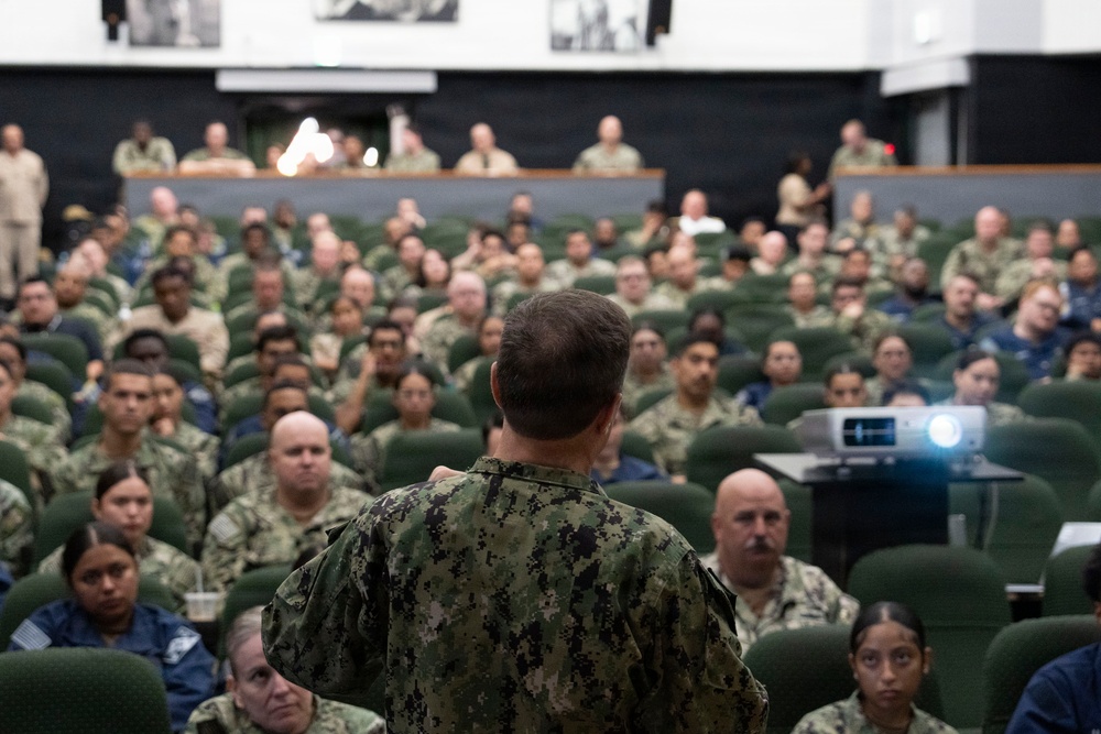 A military briefing or all-hands meeting is taking place in a large auditorium-style venue. A speaker in U.S. Navy NWU Type III (woodland digital camouflage) uniform addresses a large audience of military personnel seated in green theater-style chairs. The audience consists of predominantly Navy personnel in various camouflage uniforms, with a projection system visible to the right.
