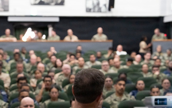 A military briefing or all-hands meeting is taking place in a large auditorium-style venue. A speaker in U.S. Navy NWU Type III (woodland digital camouflage) uniform addresses a large audience of military personnel seated in green theater-style chairs. The audience consists of predominantly Navy personnel in various camouflage uniforms, with a projection system visible to the right.