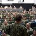 A military briefing or all-hands meeting is taking place in a large auditorium-style venue. A speaker in U.S. Navy NWU Type III (woodland digital camouflage) uniform addresses a large audience of military personnel seated in green theater-style chairs. The audience consists of predominantly Navy personnel in various camouflage uniforms, with a projection system visible to the right.