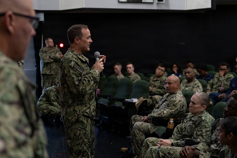 A military briefing or all-hands meeting is taking place in what appears to be an auditorium or theater space aboard a naval vessel or installation. A senior naval officer in NWU Type III (Navy Working Uniform) camouflage is addressing a seated audience of sailors using a handheld microphone. The audience consists of multiple uniformed Navy personnel seated in rows of green chairs.
