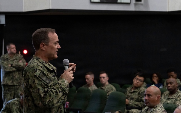A military briefing or all-hands meeting is taking place in what appears to be an auditorium or theater space aboard a naval vessel or installation. A senior naval officer in NWU Type III (Navy Working Uniform) camouflage is addressing a seated audience of sailors using a handheld microphone. The audience consists of multiple uniformed Navy personnel seated in rows of green chairs.