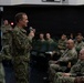 A military briefing or all-hands meeting is taking place in what appears to be an auditorium or theater space aboard a naval vessel or installation. A senior naval officer in NWU Type III (Navy Working Uniform) camouflage is addressing a seated audience of sailors using a handheld microphone. The audience consists of multiple uniformed Navy personnel seated in rows of green chairs.