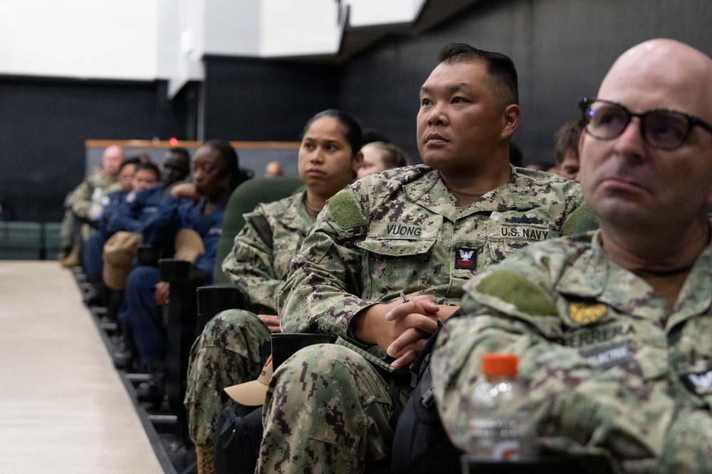 A group of U.S. Navy personnel in AOR-2 digital camouflage uniforms are seated in rows, appearing to attend a briefing, presentation, or training session. The central figure has a name tape reading 'VUONG' and 'U.S. NAVY' clearly visible, with a Petty Officer insignia on the chest. The setting appears to be an auditorium or multipurpose military facility.