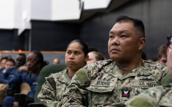 A group of U.S. Navy personnel in AOR-2 digital camouflage uniforms are seated in rows, appearing to attend a briefing, presentation, or training session. The central figure has a name tape reading 'VUONG' and 'U.S. NAVY' clearly visible, with a Petty Officer insignia on the chest. The setting appears to be an auditorium or multipurpose military facility.
