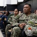 A group of U.S. Navy personnel in AOR-2 digital camouflage uniforms are seated in rows, appearing to attend a briefing, presentation, or training session. The central figure has a name tape reading 'VUONG' and 'U.S. NAVY' clearly visible, with a Petty Officer insignia on the chest. The setting appears to be an auditorium or multipurpose military facility.