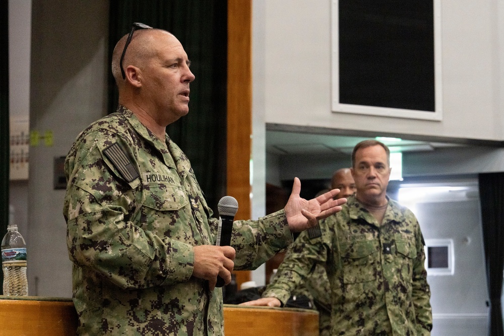 A U.S. Navy servicemember named Houlihan is speaking at a podium while holding a microphone, appearing to address an audience. He is wearing Navy Working Uniform (NWU) digital camouflage and has sunglasses resting on his head. At least two other Navy personnel in matching uniforms are visible in the background.