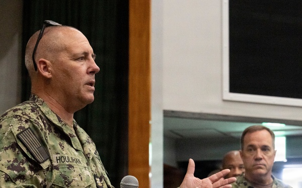 A U.S. Navy servicemember named Houlihan is speaking at a podium while holding a microphone, appearing to address an audience. He is wearing Navy Working Uniform (NWU) digital camouflage and has sunglasses resting on his head. At least two other Navy personnel in matching uniforms are visible in the background.
