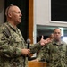 A U.S. Navy servicemember named Houlihan is speaking at a podium while holding a microphone, appearing to address an audience. He is wearing Navy Working Uniform (NWU) digital camouflage and has sunglasses resting on his head. At least two other Navy personnel in matching uniforms are visible in the background.