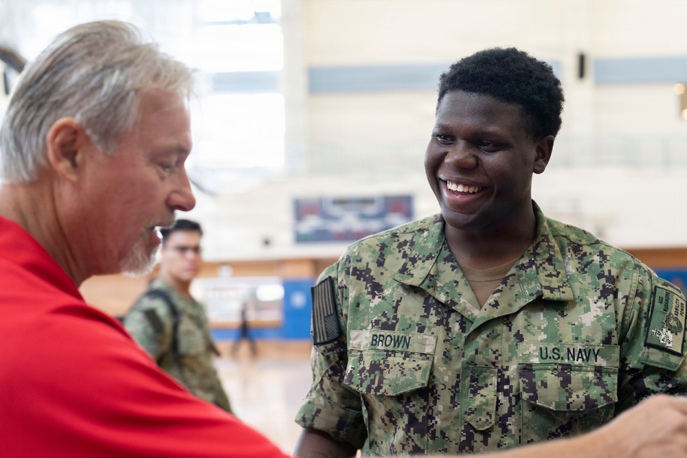 A U.S. Navy sailor named Brown is smiling broadly while interacting with an older civilian man in a red shirt, appearing to shake hands or engage in conversation. The setting appears to be an indoor gymnasium or recreational facility. Additional military personnel in camouflage uniforms are visible in the background.
