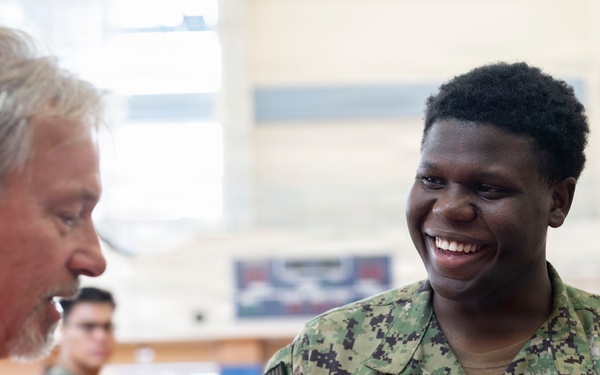 A U.S. Navy sailor named Brown is smiling broadly while interacting with an older civilian man in a red shirt, appearing to shake hands or engage in conversation. The setting appears to be an indoor gymnasium or recreational facility. Additional military personnel in camouflage uniforms are visible in the background.